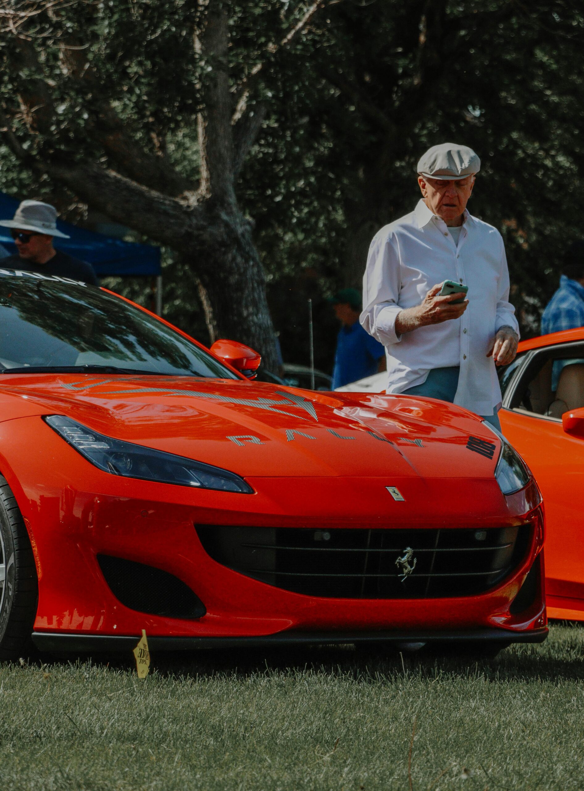 An elderly man stands beside a vibrant red Ferrari Portofino at an outdoor car show.